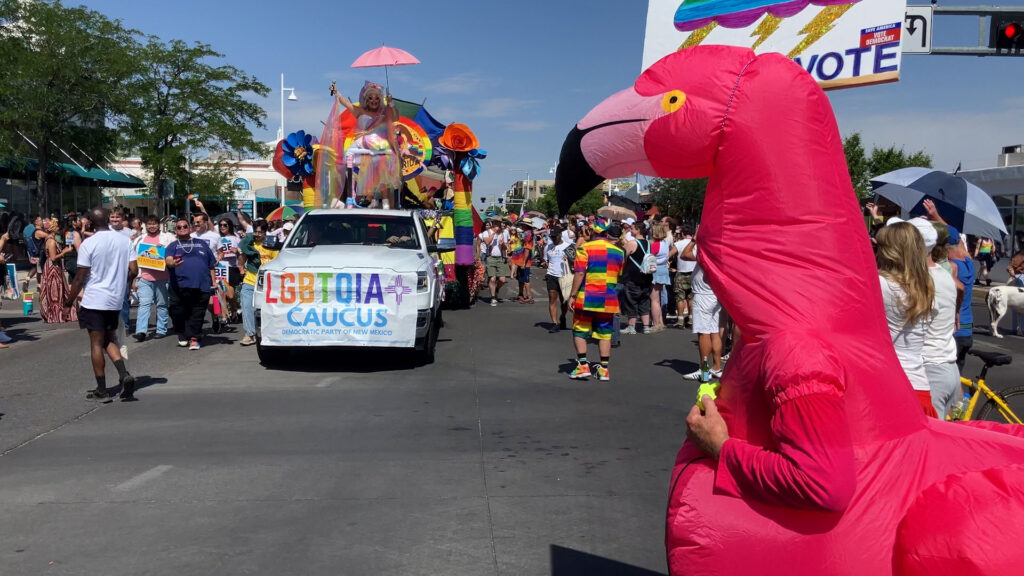 People participate in a Pride parade with a colorful float, a banner reading "LGBTQIA Caucus," and a person in a large inflatable flamingo costume.