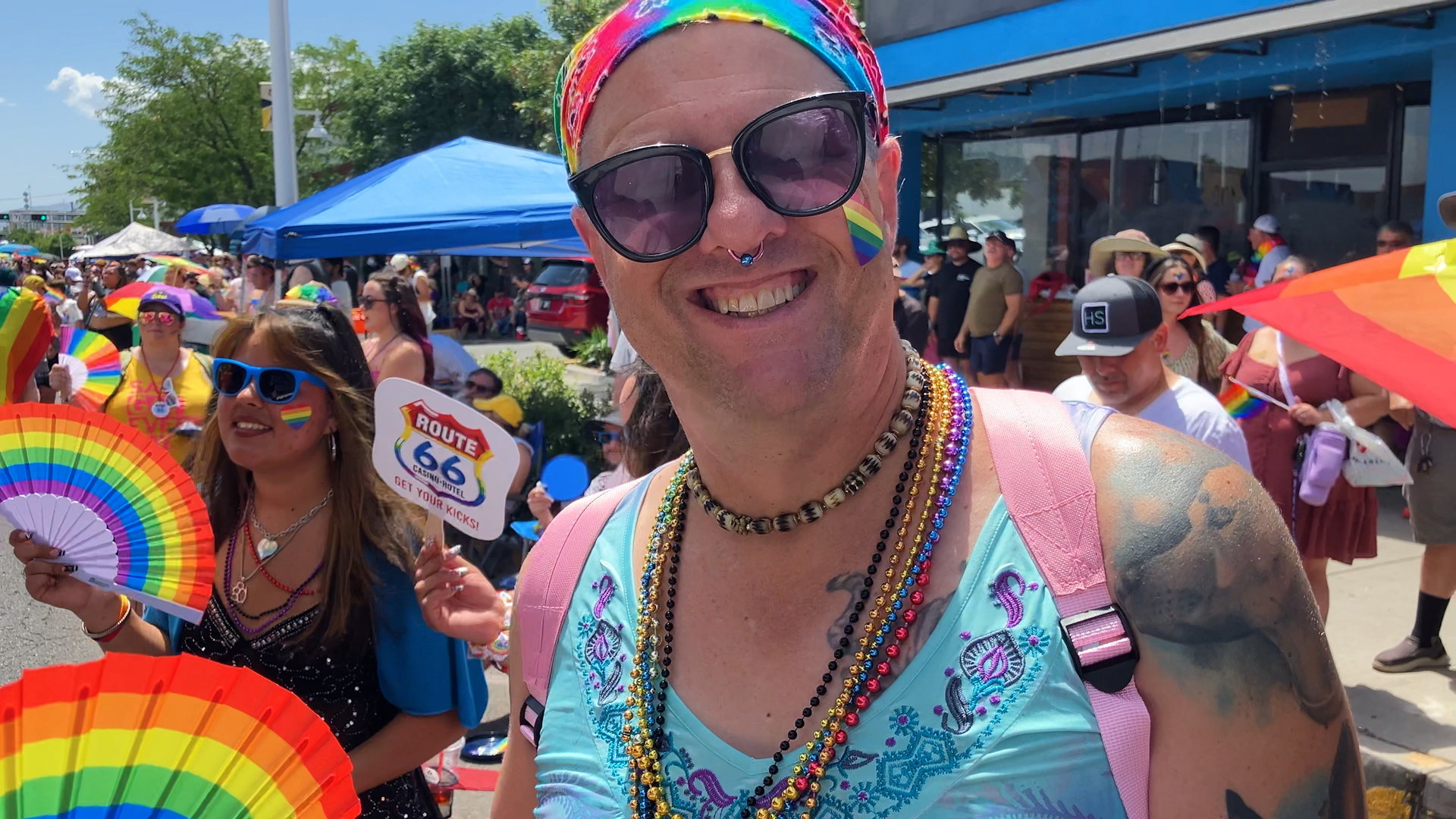 A person wearing rainbow accessories and sunglasses smiles at a pride parade, surrounded by others holding rainbow fans and flags.
