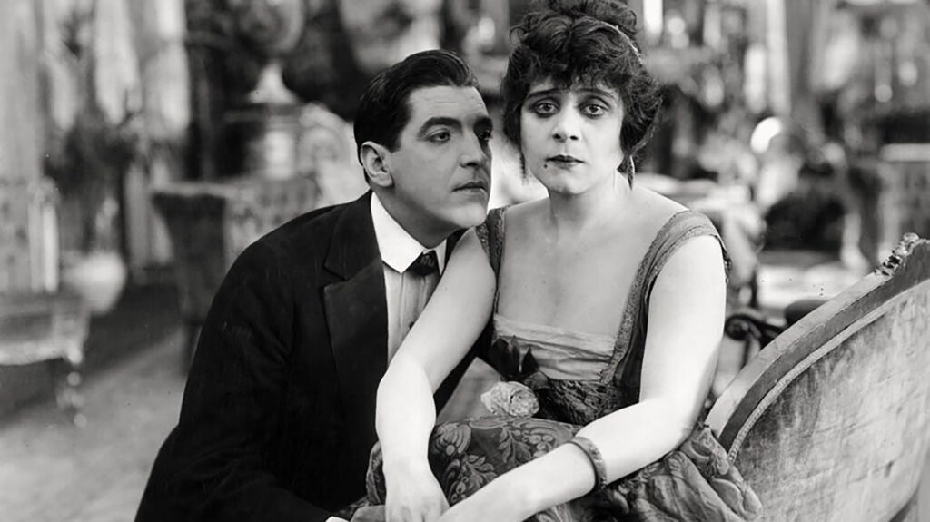 A man in a suit leans close to a woman in a dress, both sitting on a couch in a vintage, ornate room, looking intently toward the camera.