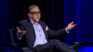 A man with glasses sits in a chair with arms outstretched, wearing a blue blazer and jeans, on a stage with dark blue lighting.