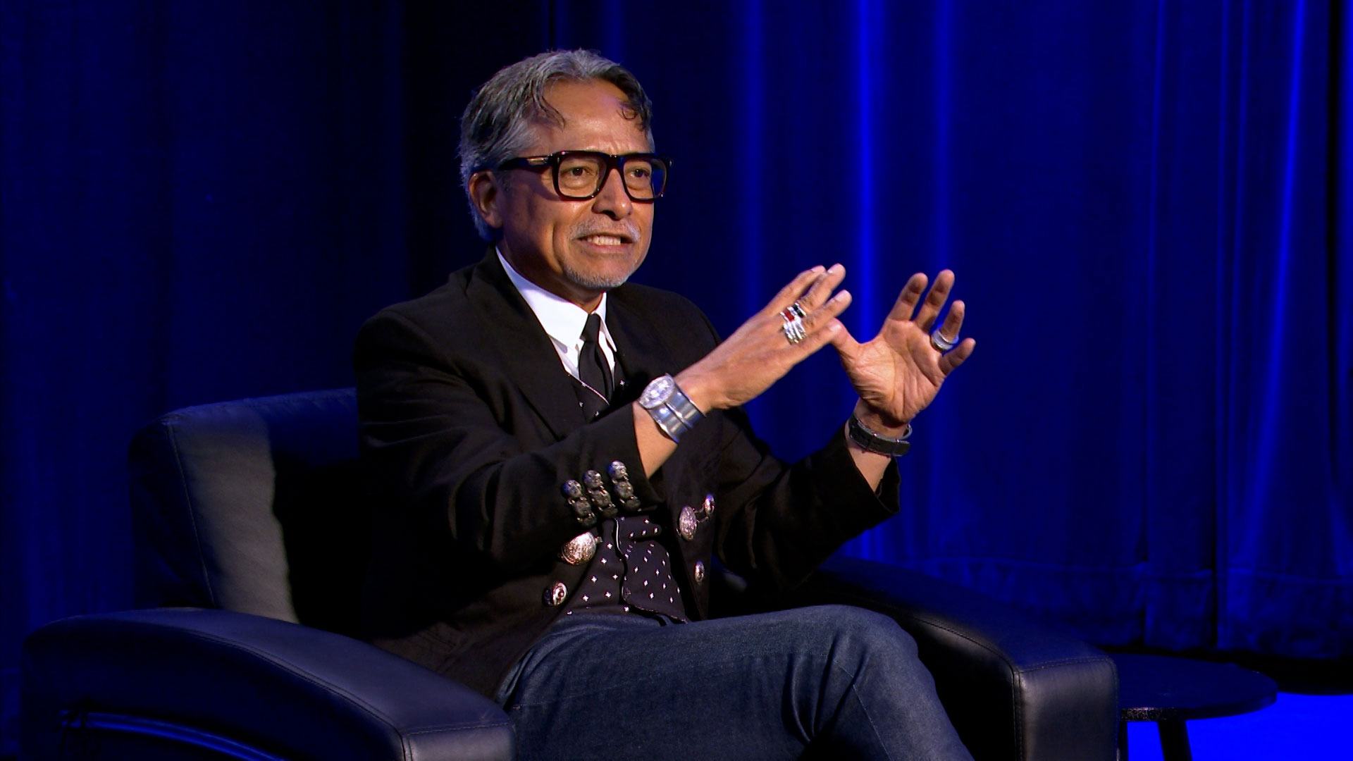 A man with glasses sits in a black chair on stage, gesturing with both hands while speaking. He wears a black jacket, patterned tie, and jeans, with a blue curtain in the background.