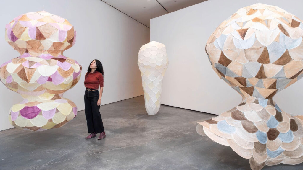 A woman stands in a gallery with three large hanging sculptures made of pastel and neutral-toned, scale-like panels.