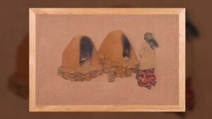 A person in a patterned skirt stands near two adobe ovens with fires inside, set on a stone base against a plain background.
