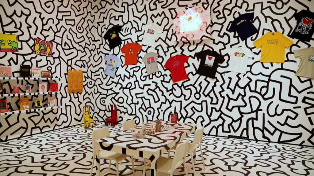 A room with black-and-white abstract line art on the walls and floor, featuring hanging t-shirts, shelves with items, and a table with chairs in the center.