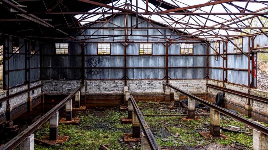 Interior of an abandoned industrial building with rusted beams, overgrown plants on the ground, and a corrugated metal wall with small windows at the far end.