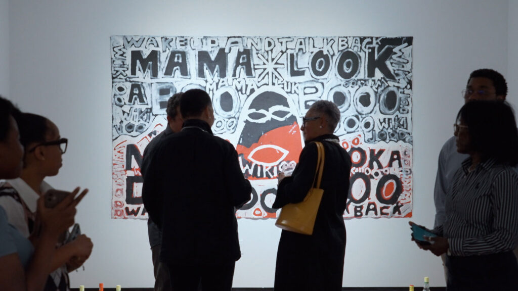 A group of people view a colorful, text-based artwork on a gallery wall that prominently displays the words “MAMA LOOK.”.