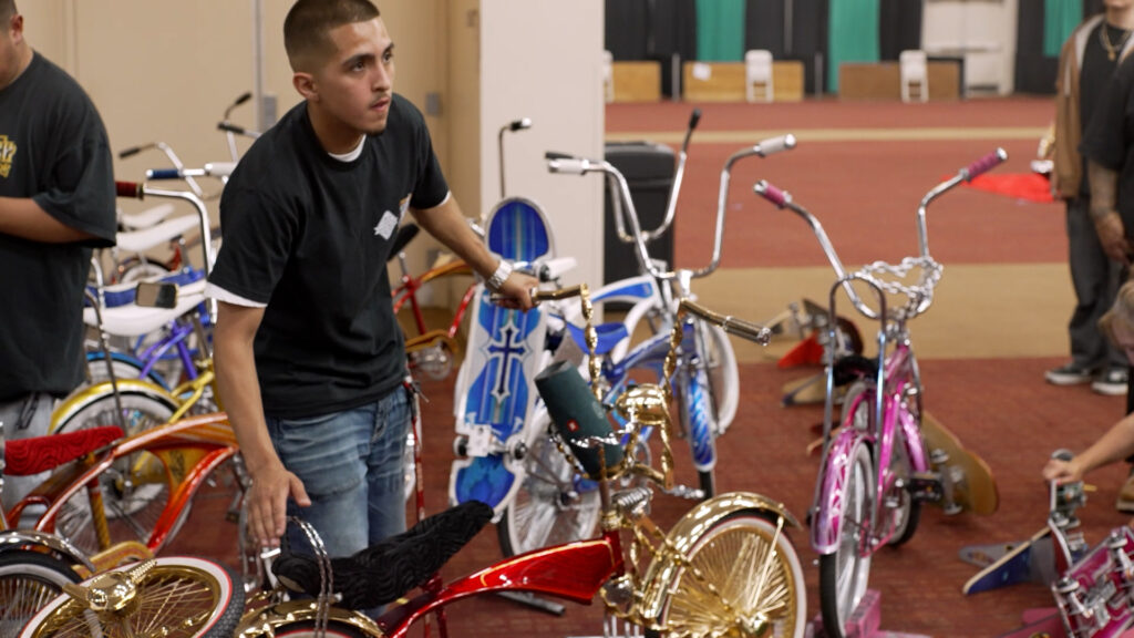 A young man stands next to a shiny red and gold lowrider bicycle at an indoor bike show, with several other custom bicycles visible nearby.