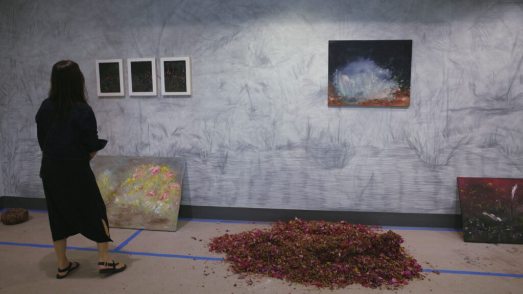 A person stands in a gallery viewing paintings on a wall; flower petals are scattered in a pile on the floor beneath the artwork.