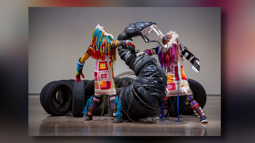 Two figures in colorful outfits and masks interact with a large, black, sculptural structure made of tires and other materials in a gallery setting.