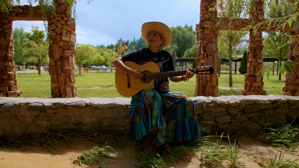 Person wearing a hat and colorful skirt sits on a stone wall outdoors, playing an acoustic guitar, with trees and grass visible in the background.