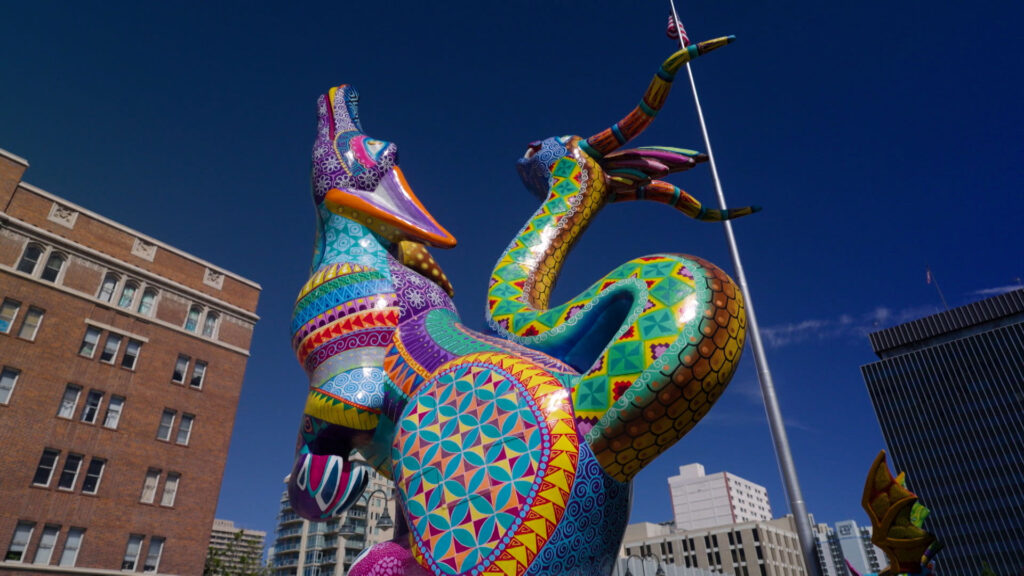Colorful, patterned alebrije sculpture of a fantastical dragon-like creature stands outdoors in an urban setting with buildings and a flagpole in the background.