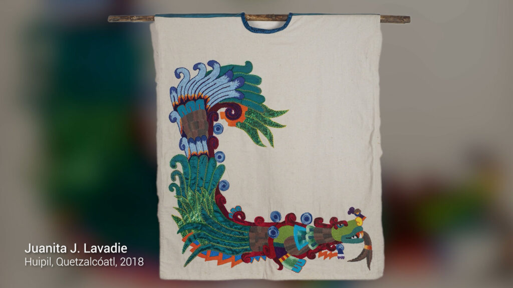 A handmade huipil featuring a colorful embroidered feathered serpent, displayed on a wooden rod. Text reads: “Juanita J. Lavadie, Huipil, Quetzalcóatl, 2018.”.