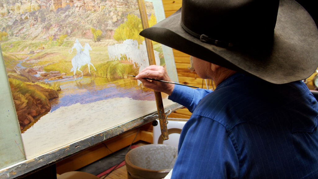 An artist in a blue shirt and black hat paints a western landscape scene with horses and cattle on a large canvas in a studio.