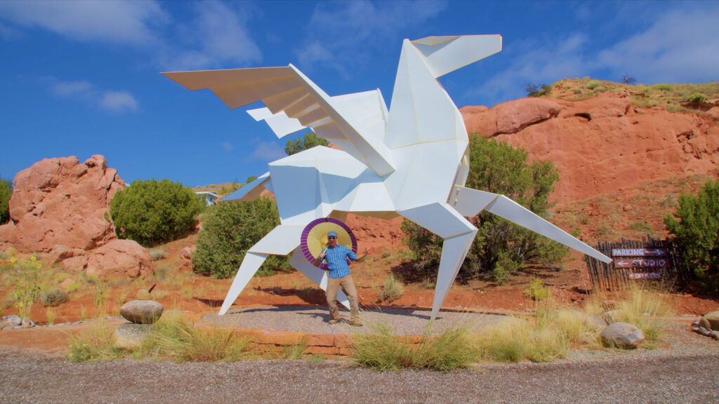 A person holding a parasol stands in front of a large, white, angular sculpture of a winged horse, with red rock formations in the background.