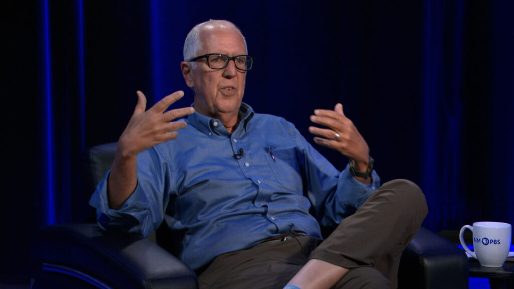 An older man in glasses and a blue button-down shirt sits in a chair and gestures while speaking.