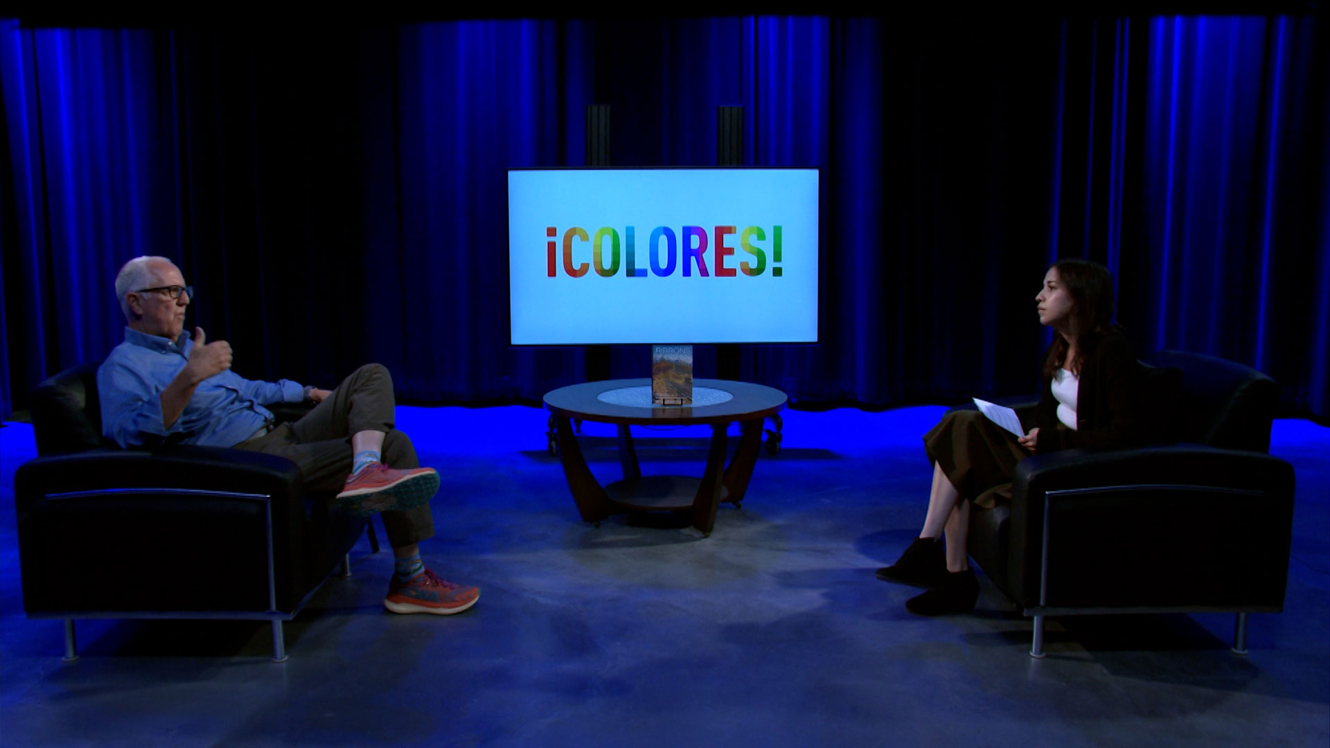 A man and woman sit in armchairs on a studio set facing a screen that reads “¡COLORES!” with a book placed on a coffee table between them.
