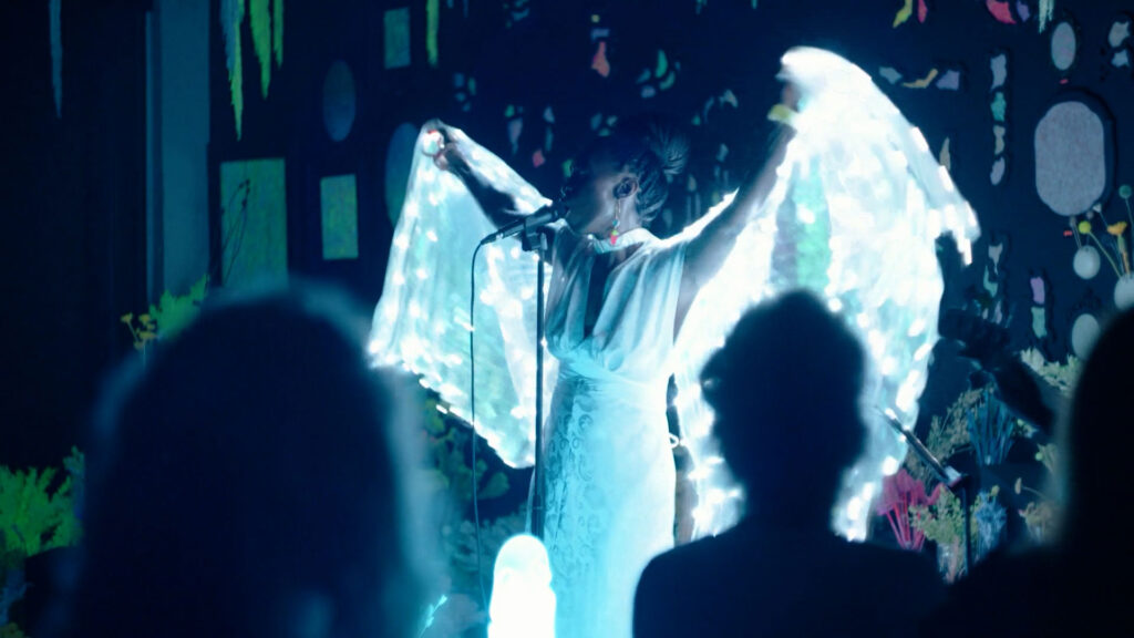 A performer in a glowing, winged costume sings onstage, arms raised, with a dark background and an audience in silhouette.