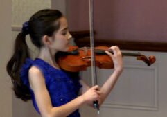 A girl in a blue dress plays the violin indoors, focusing on her performance.