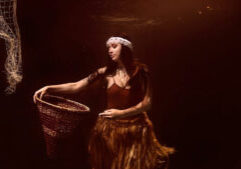 A woman underwater holds a woven basket, wearing a feather headband and a fringed skirt, with fishing nets visible in the background.