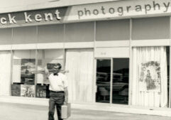 A man carrying a suitcase and equipment walks past the storefront of "dick kent photography," which has large windows displaying photos and a poster.