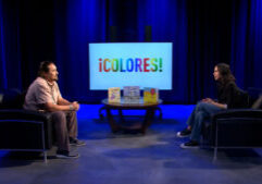 Two people sit across from each other in a studio with blue lighting. A table with books and a screen displaying "¡COLORES!" are between them.