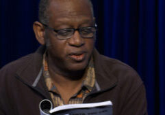 Man with glasses reading a book in front of a blue curtain.