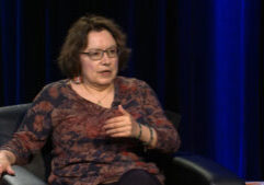 DemtriaMartinez_still003 A woman with glasses and shoulder-length hair sits in a chair, gesturing with her hand while speaking against a dark blue curtain background.