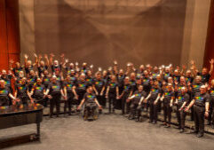 A large choir of diverse adults, some in wheelchairs, pose on stage wearing matching black shirts with rainbow lettering, arms raised, in front of a grand piano and wood-paneled background.
