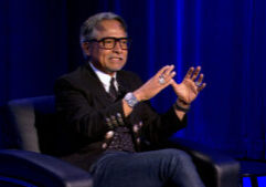 A man with glasses sits in a black chair on stage, gesturing with both hands while speaking. He wears a black jacket, patterned tie, and jeans, with a blue curtain in the background.