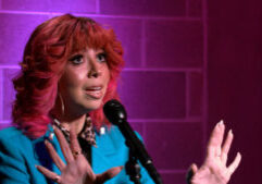 A person with pink hair in a blue jacket speaks into a microphone, gesturing with raised hands against a purple-lit brick wall background.