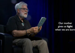 An older man with gray hair and glasses sits in a chair reading from a book. Text on screen reads: "Our mother gives us light when we are born.