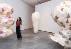 A woman stands in a gallery with three large hanging sculptures made of pastel and neutral-toned, scale-like panels.
