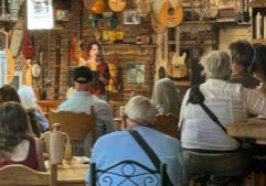 A woman stands at a microphone reading to an audience seated in a cozy, guitar-filled room with eclectic decorations and wooden furniture.