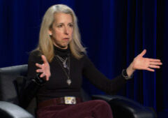 A woman with long blonde hair sits in a black chair, wearing a black turtleneck and maroon pants, gesturing with both hands in front of a dark blue curtain.
