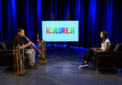 A musician plays wooden flutes while seated across from a woman in a studio with a screen displaying the word “¡COLORES!”.