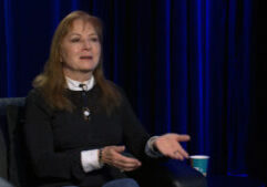 A woman with long, light brown hair sits in a black chair, gesturing with her hands, with a blue curtain and a paper cup visible in the background.