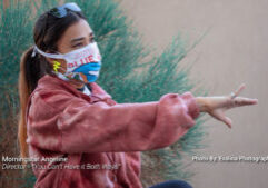 A woman in a pink jacket and colorful face mask gestures with her hand while seated outdoors near green bushes. Text on image credits Morningstar Angeline and photographer Evalina.