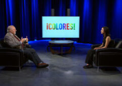 Two people sit in armchairs on a studio set with a screen displaying the word “¡COLORES!” in colorful letters.