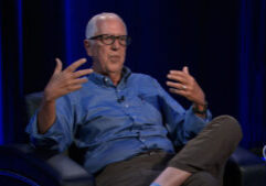 An older man in glasses and a blue button-down shirt sits in a chair and gestures while speaking.