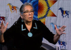 An older woman wearing glasses and turquoise jewelry speaks, gesturing with her hands. A colorful star quilt with horse designs hangs in the background.