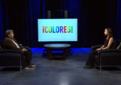 Two people sit facing each other in a studio with a round table between them and a screen displaying the word "¡COLORES!" in colorful letters.