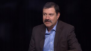 A man with a mustache sitting in front of a black background.