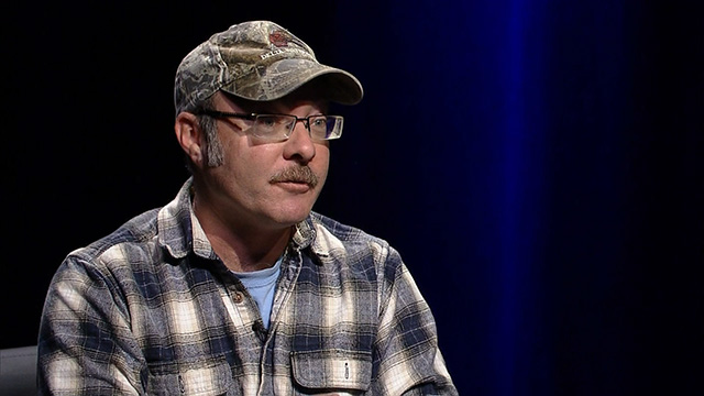 A man wearing glasses and a plaid shirt sits in front of a blue screen.