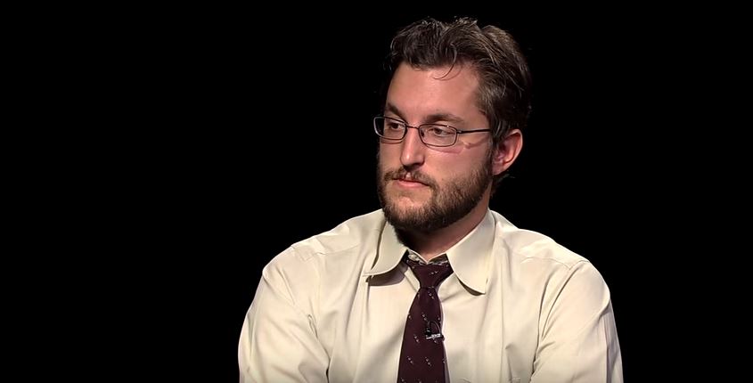 A man with glasses and a tie sitting on a black background.