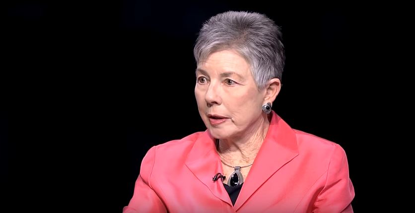 A woman in a pink jacket is talking on a black background.