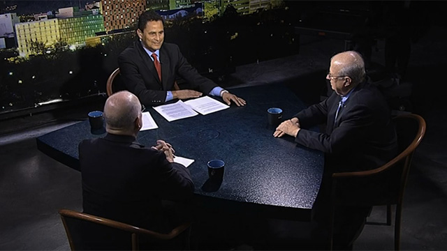 Three men sitting at a table in front of a television.