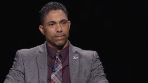 A man in a suit and tie sits in front of a black background.