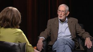 An older man sitting in a chair talking to an older woman.