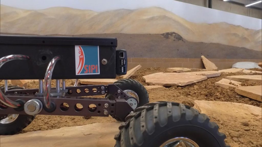 A small rover in a field with rocks in the background.