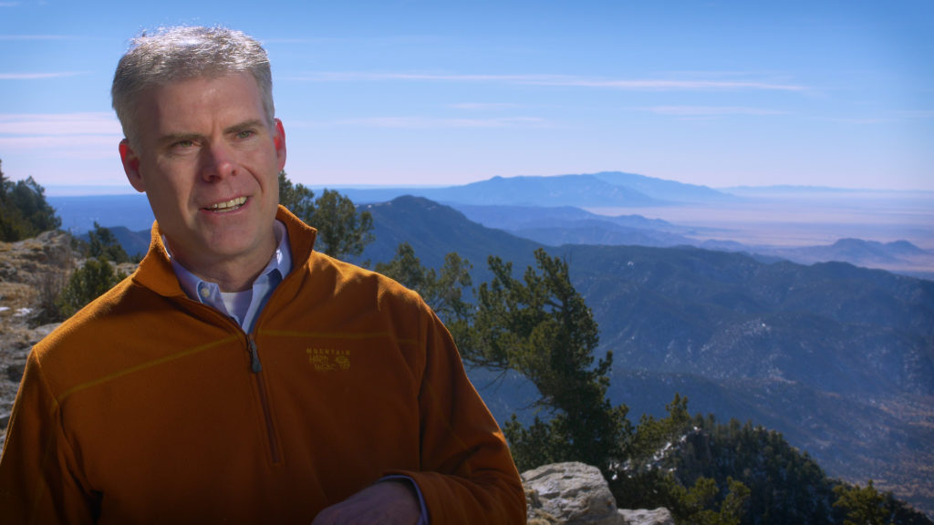 A man in an orange jacket standing on top of a mountain.